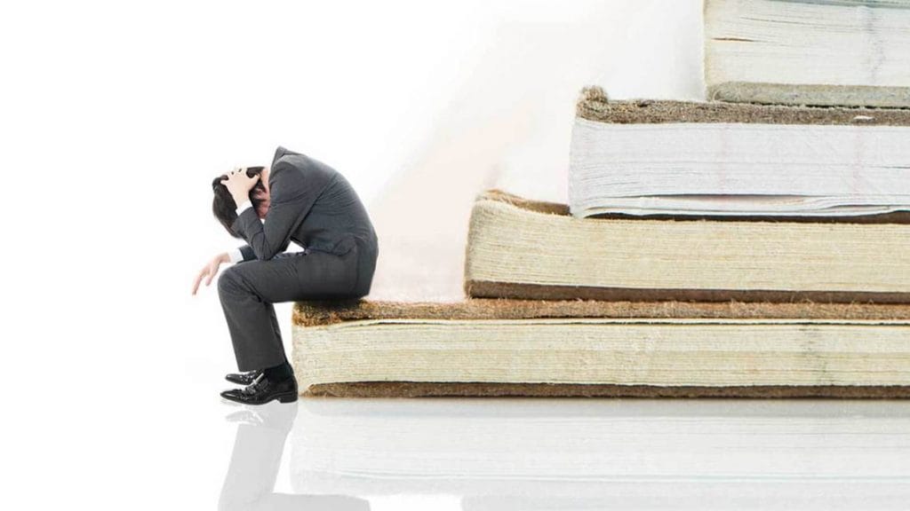 積み重なった本の上で頭を抱える悩むビジネスマン